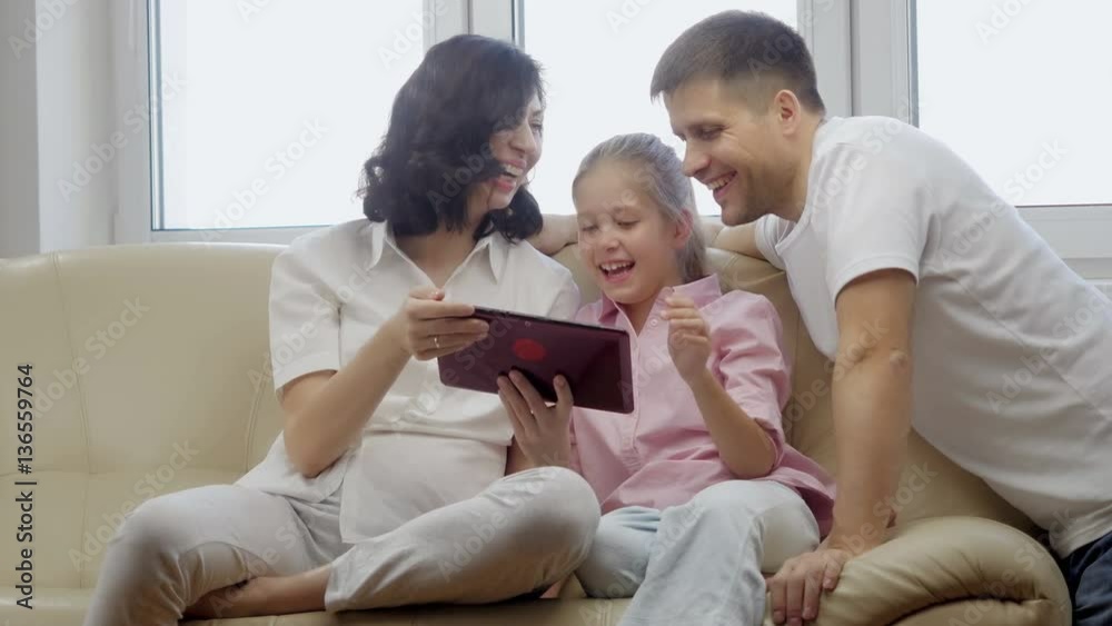 Parents showing digital tablet to their daughter at home.
Parents showing digital tablet to their daughter at home.