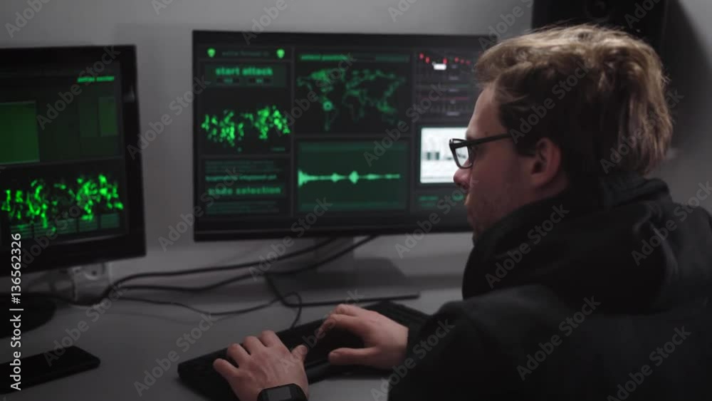 Serious man with glasses, wearing a black jacket with hood sits at the computer, and is developing a new program for government. Young programmer focused on carrying out its task