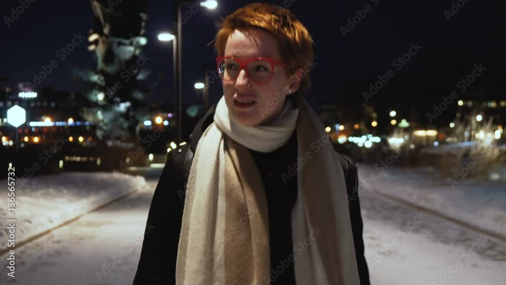 portrait of young smiling red haired woman with glasses walking in the night lighted park during cold winter evening portrait of young smiling red haired woman with glasses walking in the night lighted park during cold winter evening