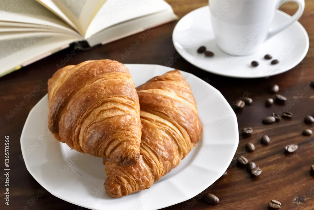 Breakfast scene with fresh croissants, coffee cup, coffee beans and open book. Relaxation morning scene with hot coffee. Breakfast scene with fresh croissants, coffee cup, coffee beans and open book. Relaxation morning scene with hot coffee.