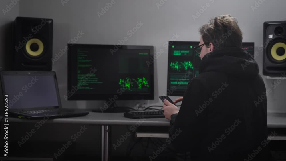 Back view of a man sitting at the table using his smartphone. Cyber spy hacking computer system with the help of his mobile phone. Hacker trying to match password and gain unauthorized access to data. Back view of a man sitting at the table using his smartphone. Cyber spy hacking computer system with the help of his mobile phone. Hacker trying to match password and gain unauthorized access to data.