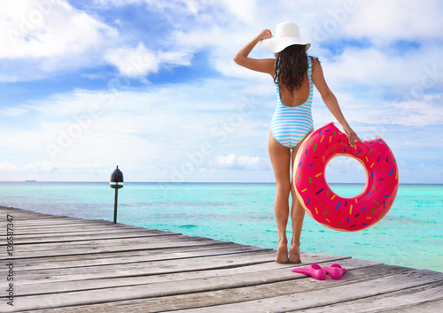 Tropical summer holiday concept, woman wearing hat and swimsuit standing on wooden pier by the beach