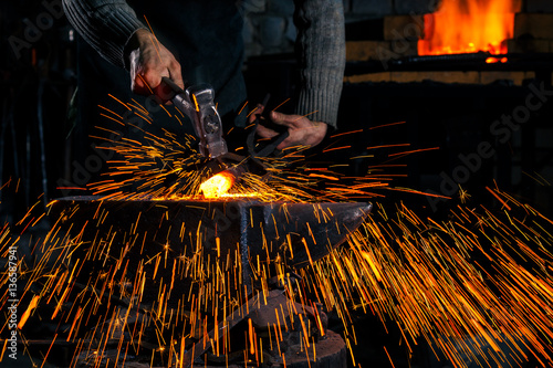 The blacksmith manually forging the molten metal on the anvil in smithy with spark fireworks