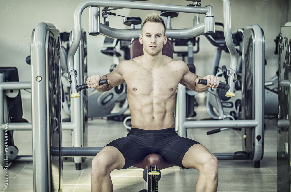 Fototapeta premium Muscular, shirtless young man in gym exercising pecs on pectoral machine