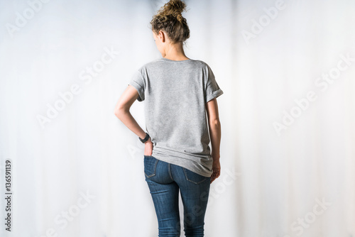 Rear view of young woman, dressed in light gray T-shirt and blue jeans, standing for light background with his hand in back pocket of jeans. Mock up. Space for text, advertising.