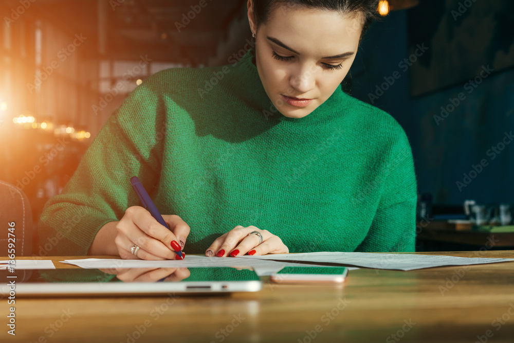 Fototapeta premium Front view. Young businesswoman sitting at table and fills out an application. On table is smartphone and tablet computer. Student learning online. Freelancer working outside office. Lifestyle.