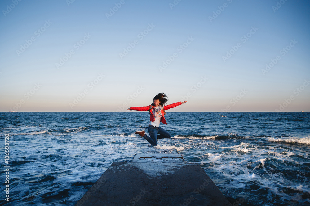 Young happy girl with backpack jumping on a seaside at sunset
