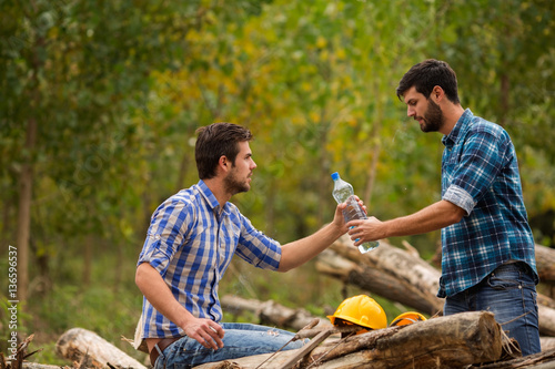 Tired young woodcutter gives bottle of water to his colleague © BanjaninFoto