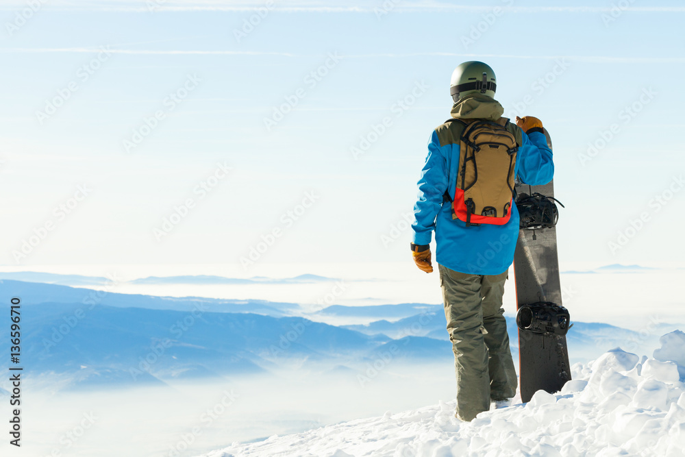 Snowboarder in helmet standing at the very top of a mountain and holding his snowboard with one hand