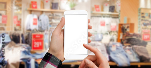 Women holding smartphone with empty screen in clothing store, touching screen with finger. Searching and shopping online, technology and fashion concept.