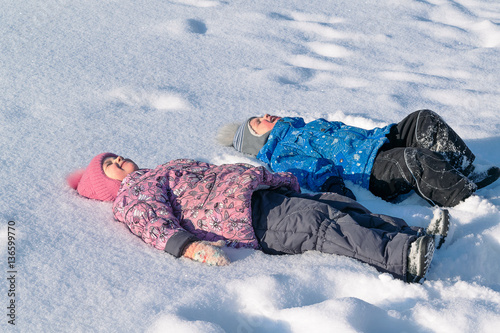 Children, brother and sister lying in the snow on the back. Laugh and smile. Dressed in jackets, hats, warm pants, boots, boots and mittens. Wear pink, blue, grey and black. He closed his eyes.