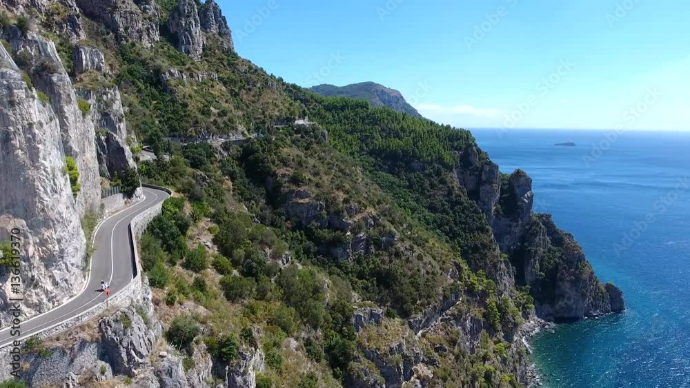 Aerial flying past beautiful coastline motorcycle driving over cliff road towards drone amazing Mediterranean landscape and motorbike driver maneuvering flexible on curvy ocean road summer vacation 4k