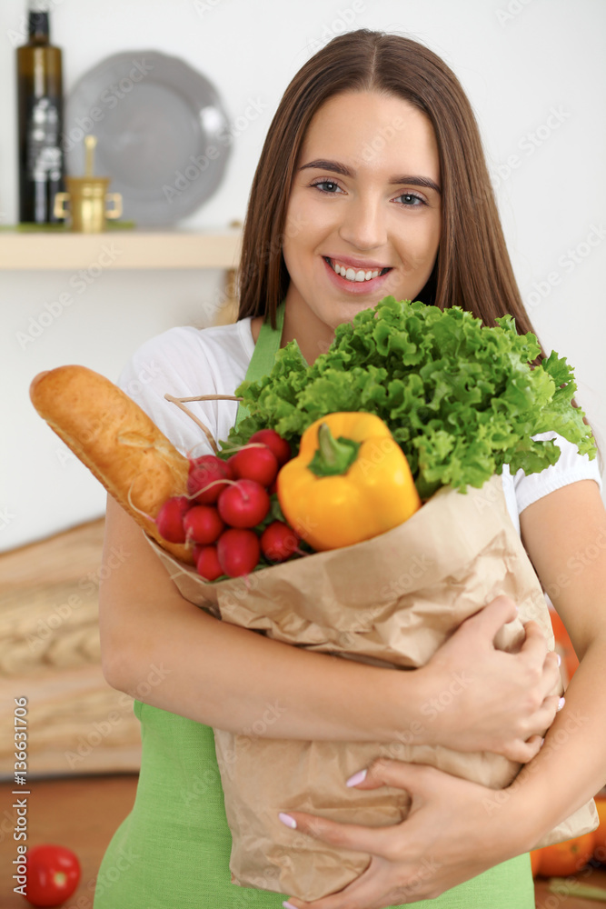 Young caucasian woman in a green apron is holding paper bag full of vegetables and fruits while smiling in kitchen Young caucasian woman in a green apron is holding paper bag full of vegetables and fruits while smiling in kitchen