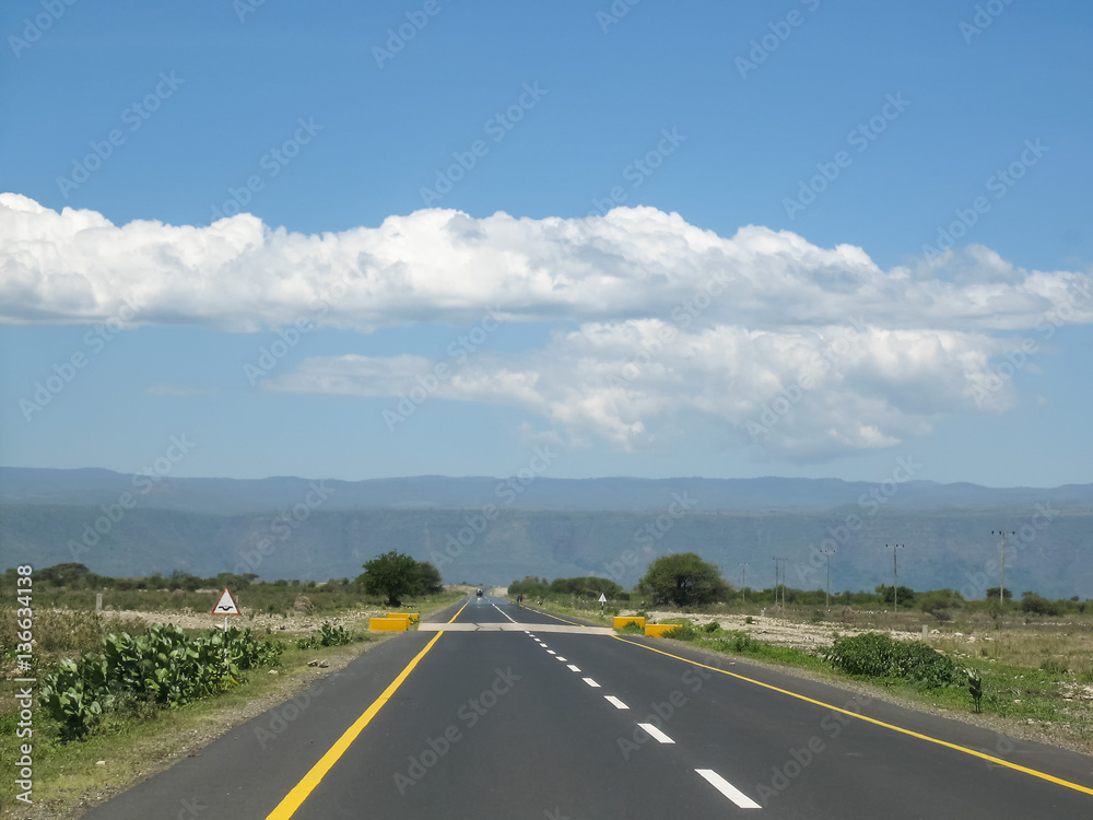 Straight modern highway in savanna vanishing in horizon against mountain and blue sky background at sunny day. Tanzania, Africa.

