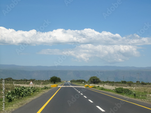 Straight modern highway in savanna vanishing in horizon against mountain and blue sky background at sunny day. Tanzania, Africa.
