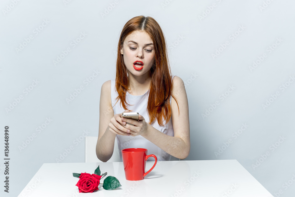 beautiful woman in bright clothes looks at the phone and a cup with a rose on the table beautiful woman in bright clothes looks at the phone and a cup with a rose on the table