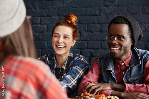 People, friendship and leisure concept. Happy interracial couple having fun at modern coffee shop, talking to their female friend and laughing cheerfully, enjoying lively and easy conversation