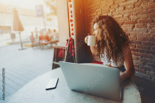 Beautiful curly brunette girl drinks tea or coffee while sitting with portable laptop in street cafe, charming dreamy woman using her computer during rest in cafe with cup of tea photo