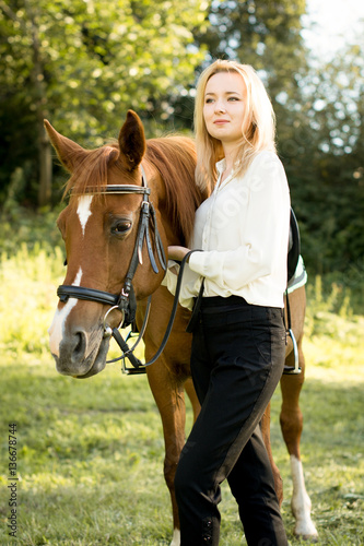 Молодая девушка со светлыми волосами стоит рядом с коричневой лошадью 