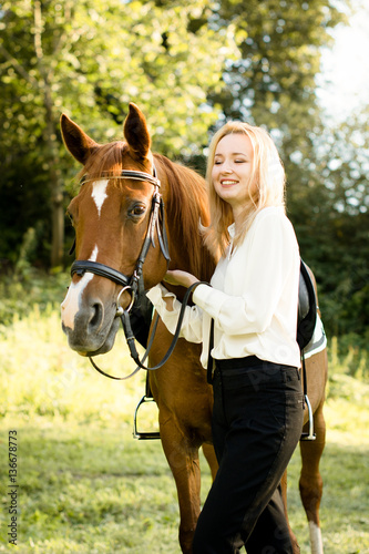 Молодая девушка со светлыми волосами стоит рядом с коричневой лошадью 