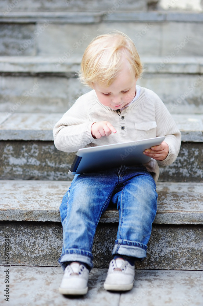 Cute blonde toddler boy playing with a digital tablet outdoors Cute blonde toddler boy playing with a digital tablet outdoors
