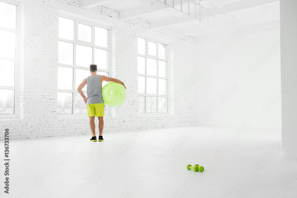 Wide shot of the white gym interior with big windows and man standing with fitball Wide shot of the white gym interior with big windows and man standing with fitball