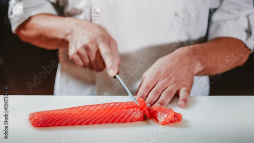 Chef coock dressed white uniform cut fresh red salmon fish with sharp knife on the cutting board in restaurant. He is working on sashimi. Preparing traditional japanese sushi set. Only hands close up