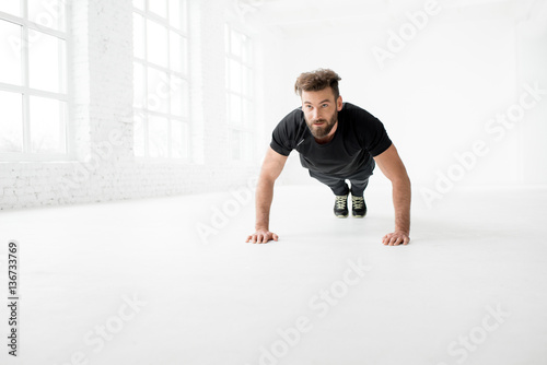 Handsome man in the black sportswear making pushups indoors in the white gym interior