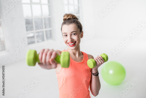 Portrait of young woman in sportswear with dumbbells and fitness ball in the white gym