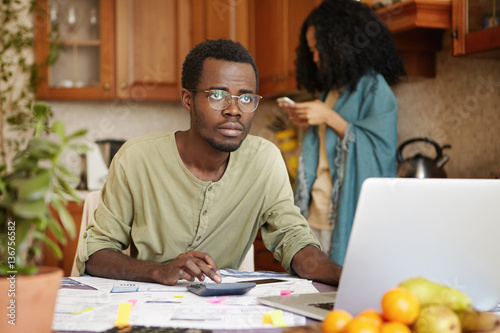 Sad unemployed African male wearing glasses having stressed look, realizing that he is not able to pay gas and electricity bills anymore, calculating family budget using laptop and calculator