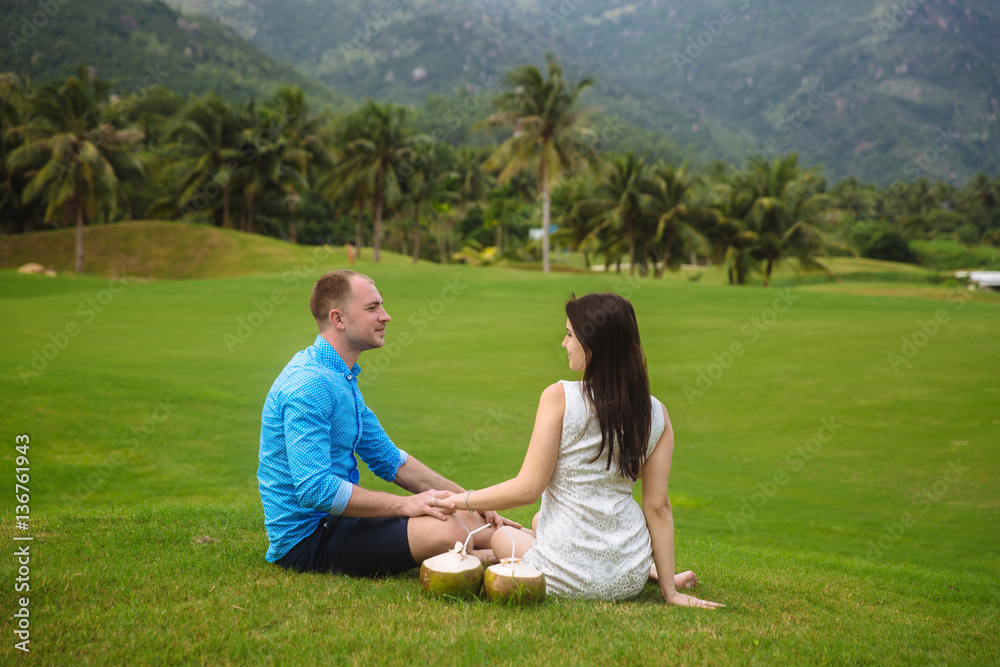 Young beautiful couple with coconuts holding their hands at golf course in summer. man wear the blue shirt and the girl in a white dress. Concept of honeymoon Young beautiful couple with coconuts holding their hands at golf course in summer. man wear the blue shirt and the girl in a white dress. Concept of honeymoon