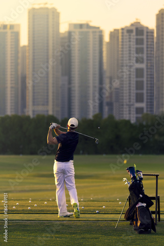 Golf player in a dark shirt and white pants hitting golf ball towards skyscrapers. Golf bags and sticks on the right side and buildings on the background