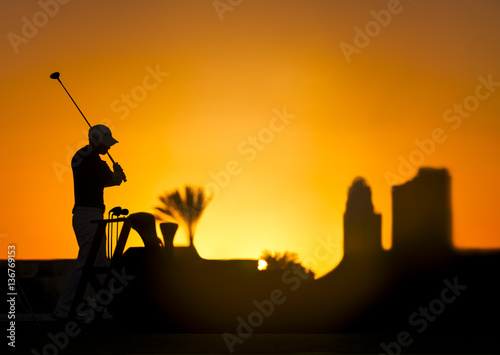 Golf player silhouette on a colorful sunset holding a golf stick. Buildings, sun and palm tree on the background.