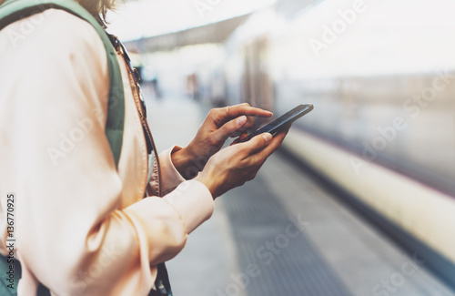 Enjoying travel. Young pretty woman waiting on the station platform with backpack on background electric train using smartphone. Tourist texting message and plan route of railway, railroad transport