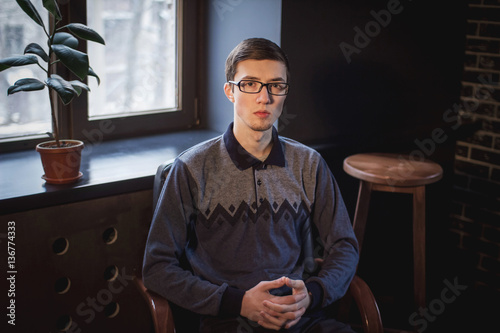 Portrait of handsome serious young man, wearing glasses, sitting in a dark room near the window