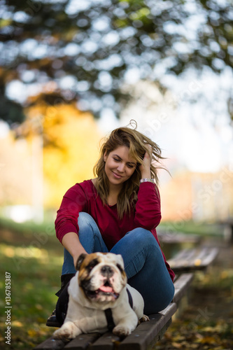 People and dogs outdoors. Portrait of beautiful and happy woman enjoying in autumn park. She sitting on bench with her adorable English bulldog.