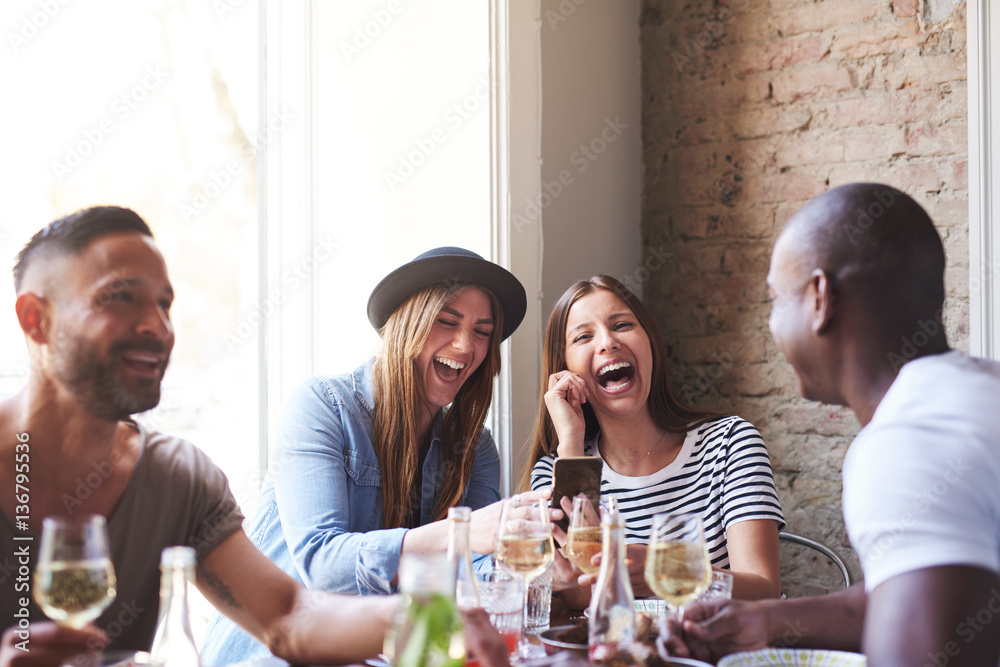 Friends laughing at something on phone at dinner - obrazy, fototapety, plakaty 