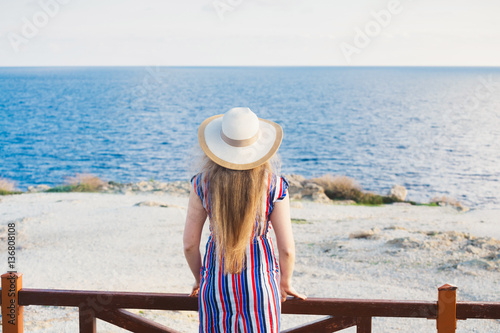 Back view of woman standing in summer dress and hat looking out towards blue ocean and sky