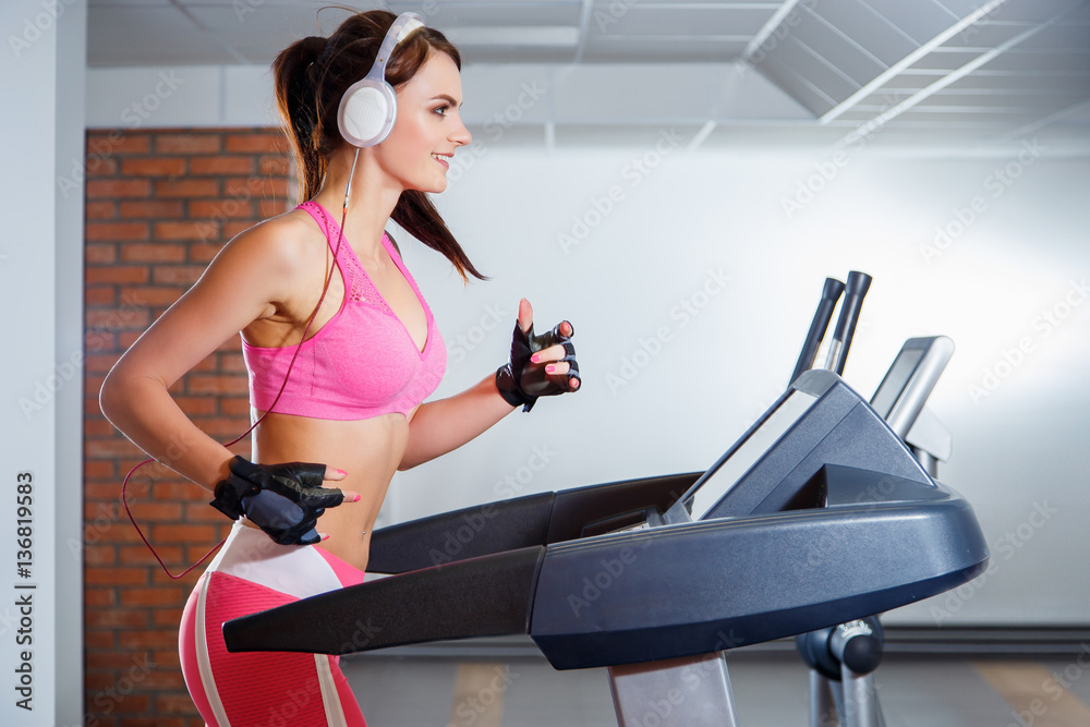 Fototapeta premium Young smiling girl with headphones running on a treadmill in a sport club. The concept of sport and active lifestyles.
