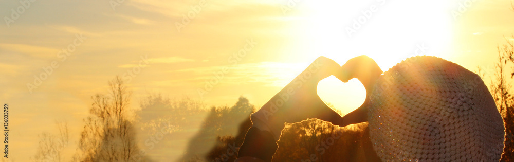 Fototapeta premium Heart Symbol. Woman hands in gloves heart symbol shaped, on a beautiful sunset light background.