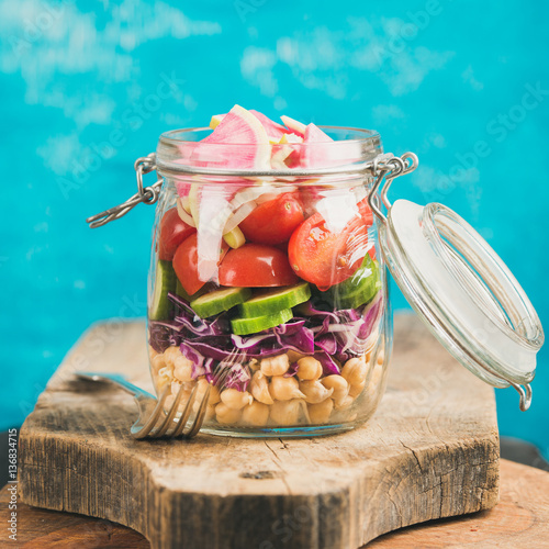 Healthy take-away lunch jar. Vegetable and chickpea sprout vegan salad in jar on wooden board, bright blue wall at background, selective focus, square crop. Clean eating, vegetarian, dieting concept