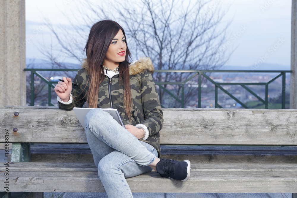 Young beautiful woman sitting on a park bench, outdoors at sunset, thinking and writing something in her notebook with a black pen. Young beautiful woman sitting on a park bench, outdoors at sunset, thinking and writing something in her notebook with a black pen.