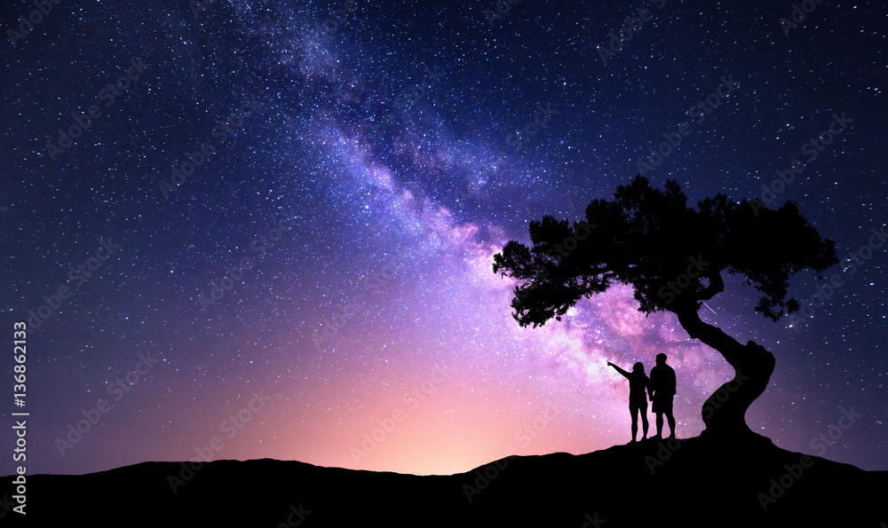 Milky Way with people under the tree on the hill. Landscape with night starry sky and silhouette of standing happy man and woman who pointing finger in starry sky. Milky way with travelers. Universe