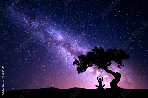 Milky Way with tree and silhouette of a sitting woman practicing yoga. Beautiful landscape with meditating girl under the tree against starry sky with purple milky way. Galaxy. Beautiful universe