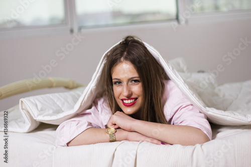 Young beautiful woman smiling under the bed covers. Relaxing at home. Woman laying in bed.