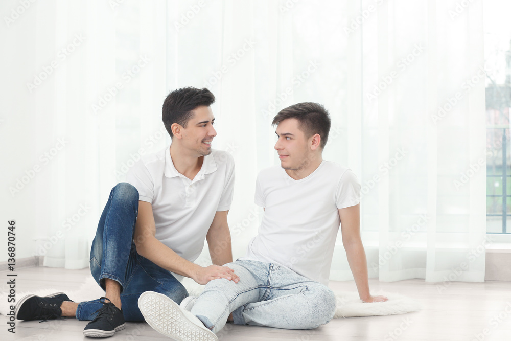 Happy gay couple sitting on floor against window Happy gay couple sitting on floor against window