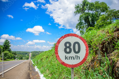 The road sign of 80 kilometres per hour limit on the background of small hill covered with green grass, long road going to horizon and blue sky with white clouds at sunny summer day