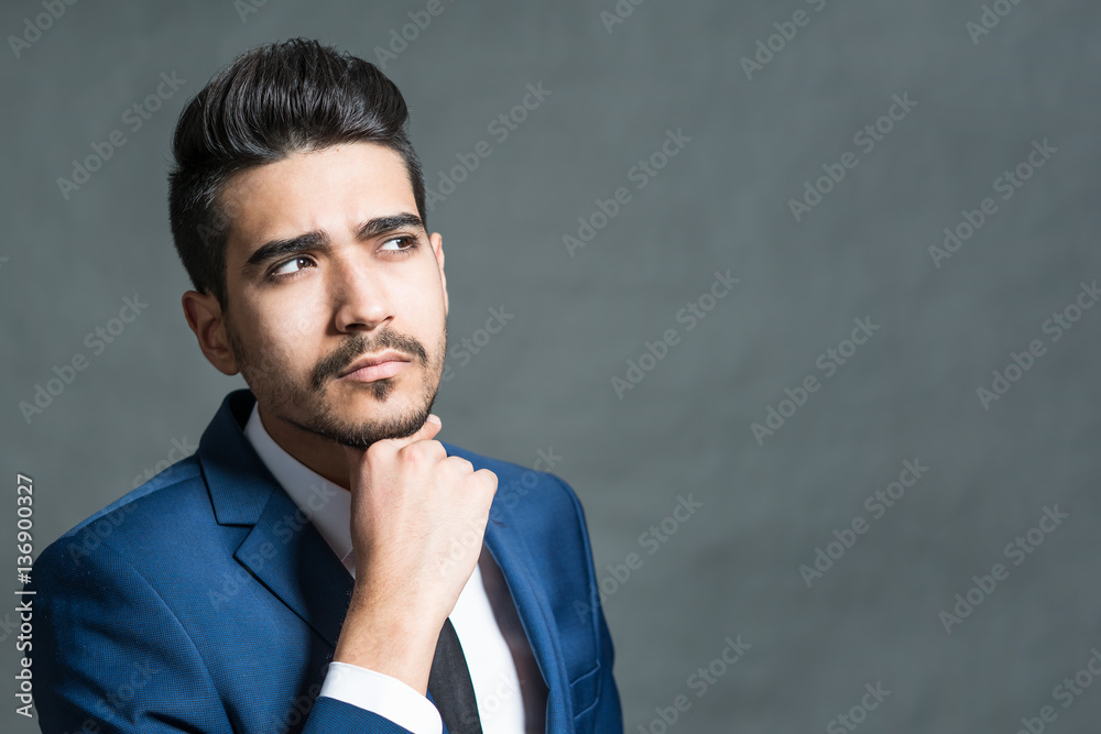 Pensive young attractive man in a blue suit with hand on chin on a gray background