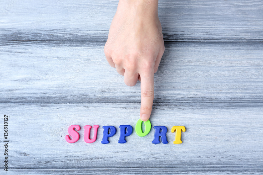 Hand adding letter O to word SUPPORT made of color plastic letters on grey wooden background Hand adding letter O to word SUPPORT made of color plastic letters on grey wooden background