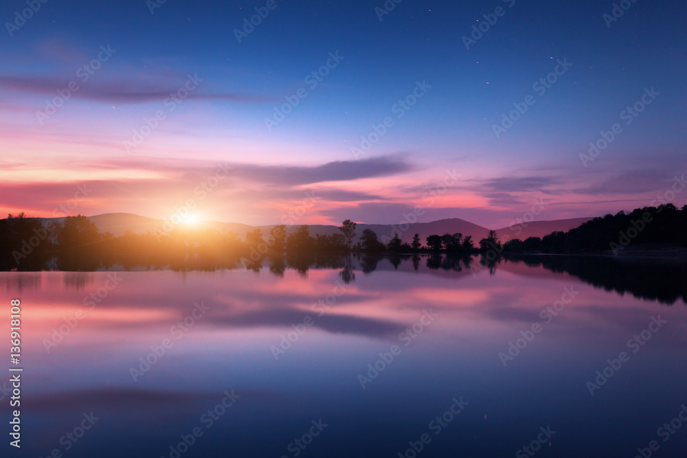 Mountain lake with moonrise at night. Night landscape with river, trees, hills, moon and colorful purple sky with clouds reflected in water in dusk. Beautiful nature background in twilight. Travel